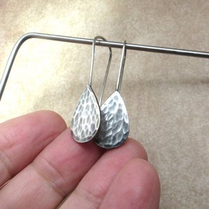 SS hammered teardrop earrings hanging from stand and displayed touching fingers.