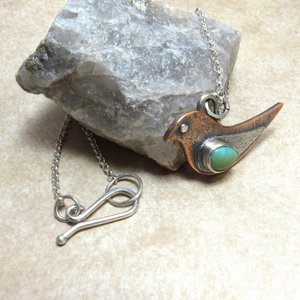 Close up of necklace hook and chain on mixed metal bird pendant set with turquoise and displayed against a rock on neutral background