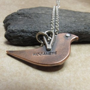 Reverse side of mixed metal copper and silver bird pendant showing our makers mark "Mocahete". Displayed against black rock on neutral background.