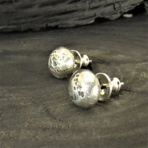 A pair of silver stud earrings with a rustic textured finish. The earrings are round and have a slightly irregular shape. Photo against dark background.