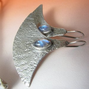 Side view of large, futuristic argentium sterling silver and rainbow moonstone earrings with a touch of rusticity. Unusual and unique earrings that are modern with hints of the past. 1.5 inches tall x 1.75 inches wide. Displayed against orange background.