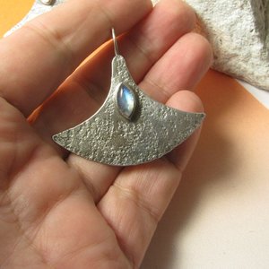 Large, futuristic argentium sterling silver and rainbow moonstone earrings with a touch of rusticity. Unusual and unique earrings that are modern with hints of the past. 1.5 inches tall x 1.75 inches wide. Displayed in hand against white rock, orange background.