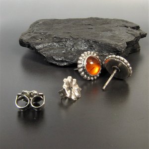 A pair of silver earrings with amber stones. The earrings are round and have a beaded edge and have SS clutch backs. The amber stones are a warm orange color. Displayed against rock on grey background.
