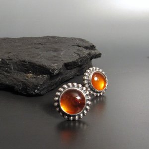 A pair of silver post back earrings with amber gemstone cabochons set in a beaded bezel. Displayed on dark rock and background