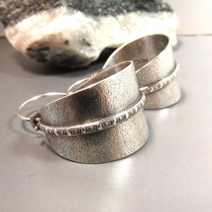 Side view of Sterling silver hoop earrings basket style fancied up with a strip of patterned  silver wire against textured argentium silver. A perfect sterling silver hoop earring for every occasion!  Oxidized, polished, finished with sterling ear wires. 1.25" or 3.1cm tall,  7/8" or 2.22cm wide. Displayed on neutral background with black and white rock.