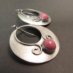 A pair of sterling silver hoop earrings with a pink rhodonite set in each. The earrings have a textured surface and a delicate design. Earrings measure a touch over 1.75” tall and a touch over 1.5” wide. Displayed against grey background.