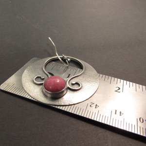 Large sterling silver earrings with earthy deep pink rhodonite cabochons, textured surface and  swirling SS wire embellishments. Oxidized to add depth and drama. Earrings measure apx 1.75” tall and a touch over 1.5” wide. Displayed against ruler on dark background.