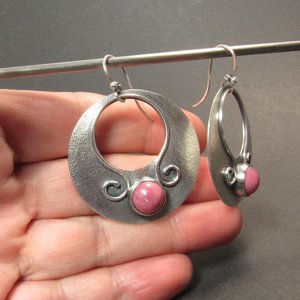 Large sterling silver earrings with earthy deep pink rhodonite cabochons, textured surface and  swirling SS wire embellishments. Oxidized to add depth and drama. Earrings measure apx 1.75” tall and a touch over 1.5” wide. Displayed against hand on dark background.