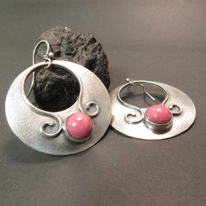 Large sterling silver earrings with earthy deep pink rhodonite cabochons, textured surface and  swirling SS wire embellishments. Oxidized to add depth and drama. Earrings measure apx 1.75” tall and a touch over 1.5” wide. Displayed on dark background.