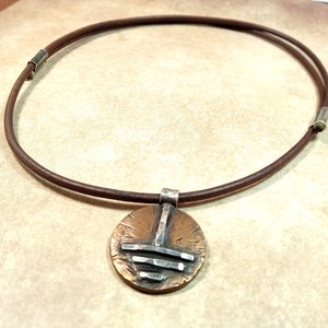 Full view of unisex bronze and silver grounding symbol pendant with adjustable leather cord, photo in hand.