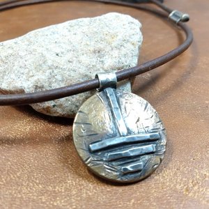 Close up of rustic unisex pendant, grounding symbol in bronze and argentium silver photo against rock.