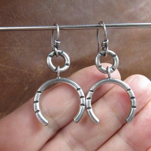 Close up of contemporary sterling silver earrings crafted from Argentium have lovely swingy movement. The double open hoop design allows for them to articulate. Lightweight and comfortable  at only 5 grams per earring. 1.75” long and 7/8” at widest point. Displayed against hand on brown leather.