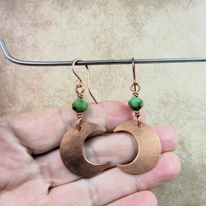 Handmade copper crescent moon dangle earrings with faceted green chrysoprase beads. Earrings measure 2 and 1/8" long and the moon is 3/8" wide at widest point.The ear wires are solid, pure copper. Displayed against hand hanging from stand.