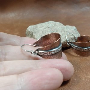 Close up of Two tone mixed metal basket shaped hoop earrings in copper and sterling silver. The textured copper hoops are encircled with a floral pattern sterling silver wire and SS ear wires. Displayed in hand on light brown back ground with white rock.