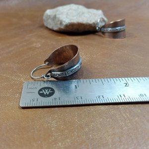 Two tone mixed metal basket shaped hoop earrings in copper and sterling silver. The textured copper hoops are encircled with a floral pattern sterling silver wire and SS ear wires. Displayed against ruler on light brown back ground with white rock.
