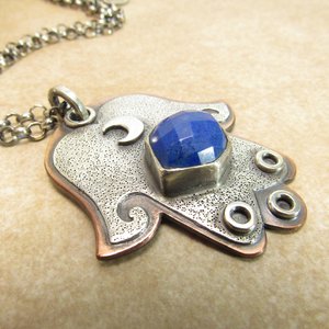 Mixed metal, copper and argentium sterling silver hamsa pendant necklace with lapis lazuli cabochon and crescent moon detail. 19 inch ss chain and findings. Hamsa is 1.5" wide x 1.75" tall. Hamsa is laying flat and displayed  on neutral background.