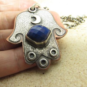 Mixed metal, copper and argentium sterling silver hamsa pendant necklace with lapis lazuli cabochon and crescent moon detail. 19 inch ss chain and findings. Hamsa is 1.5" wide x 1.75" tall. Displayed in hand on neutral background.