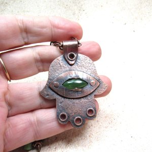 Handcrafted copper, jade and chrysoprase hamsa pendant necklace. 2" tall x 1.5" wide, 24.5" copper scroll chain with 3 faceted chrysoprase beads on each side. All riveted design. Displayed against fingers of hand on neutral background.