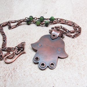 Reverse side of handcrafted copper, jade and chrysoprase hamsa pendant necklace showing makers mark "Mocahete". 2" tall x 1.5" wide, 24.5" copper scroll chain with 3 faceted chrysoprase beads on each side. All riveted design. Displayedon neutral background.