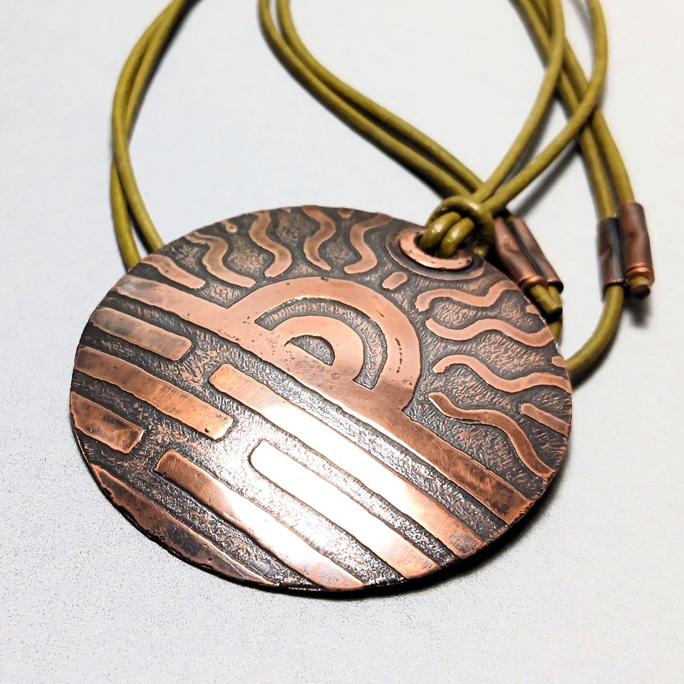 Up close of etched copper sun pendant,with adjustable cord displayed against white backgrounnd.