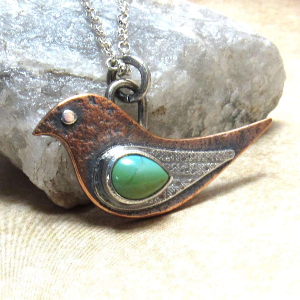 Two tone, copper and silver metal bird pendant with turquoise by Mocahete. Displayed against rock on neutral background.