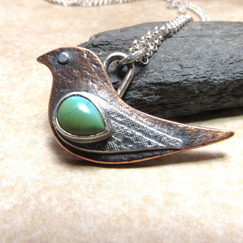 Close up of bird pendant by Mocahete, made of copper and argentium silver with a turquoise cabochon displayed against black rock on neutral background.