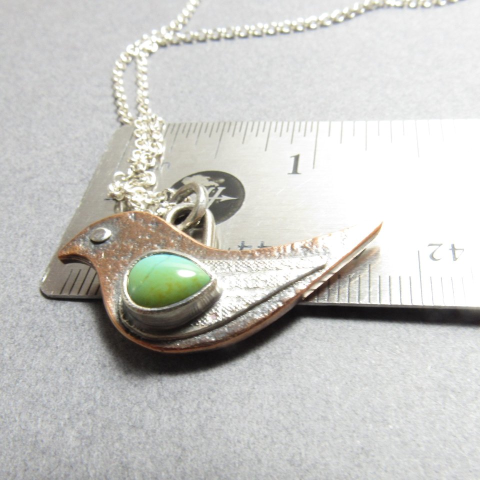 Mixed metal bird pendant necklace with turquoise displayed against ruler showing size at apx. 1.33" from beak to tail.