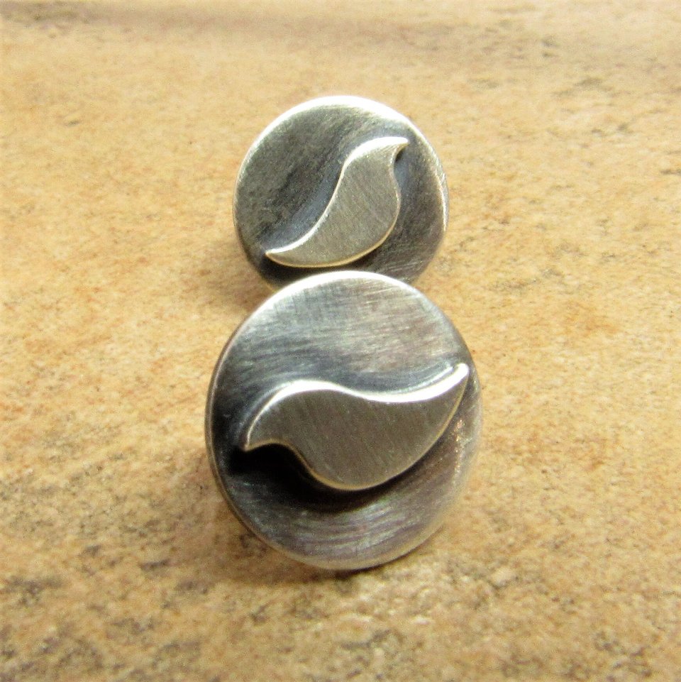 Sterling silver stud earrings with a bird design. The earrings are round and have a brushed finish displayed against sand colored tile.