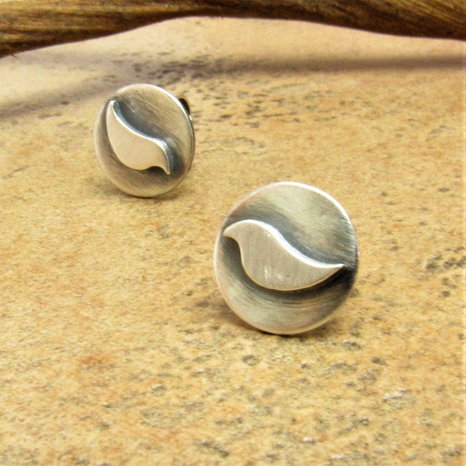 SS stud earrings with  a bird design. The earrings are round and have a brushed finish. Displayed with a twig and sand colored tile.
