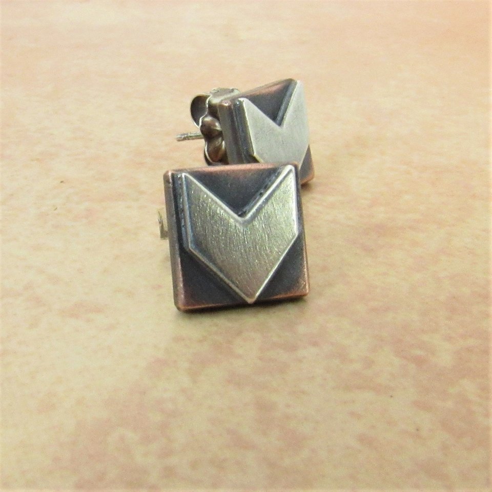 A pair of silver and copper earrings with a geometric arrow design. The earrings are square shaped with a brushed silver arrow in the center.