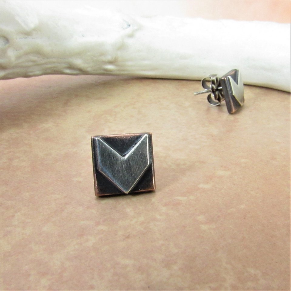 Sterling silver and copper stud earrings with a geometric arrow design. The earrings are square shaped with a pointed arrow in the center. Displayed against antler on sand color background.
