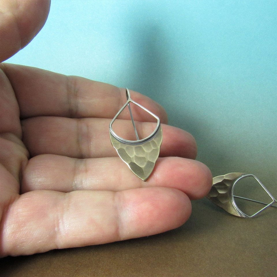 Striking, handcrafted sterling silver and bronze earrings have a two tone geometric design. Hammered for a knapped arrowhead appearance , oxidized and polished. Designed with a sterling silver fixed ear wire which holds them close to flatter your face. 1-5/8" (4.12cm) tall and 7/8" (2.2cm) wide.  Displayed in hand against blue and brown background.