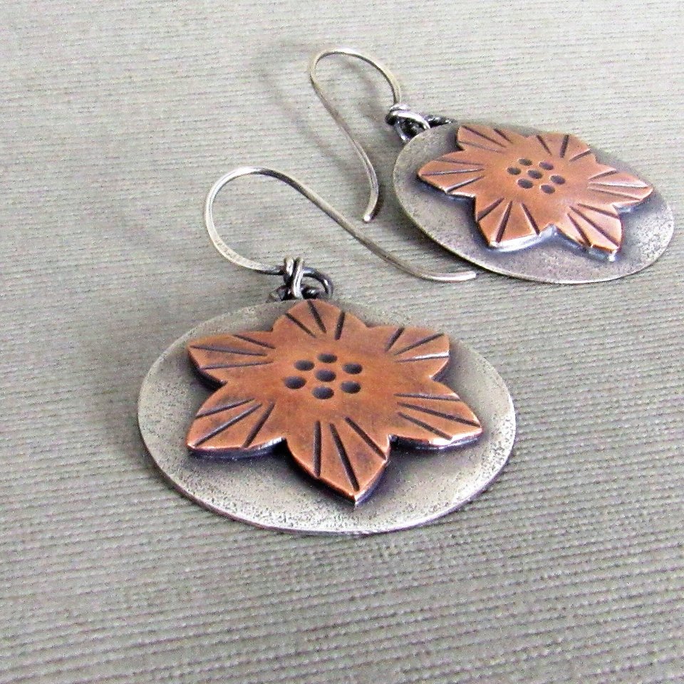 A pair of handcrafted sterling silver earrings with copper flower accents. The flowers have a textured design and are set on a round silver base. Ear wires are sterling silver. The earrings are lightweight and measure 1.5 inches tall and 1 inch wide. Displayed on grey background.