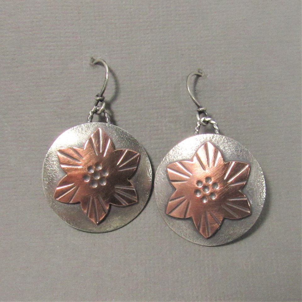 Sterling silver and copper flower earrings. The earrings are round and have a textured surface. they measure 1.5" tall and 1" wide. Handmade in the Mocahete studio. Displayed on grey background.
