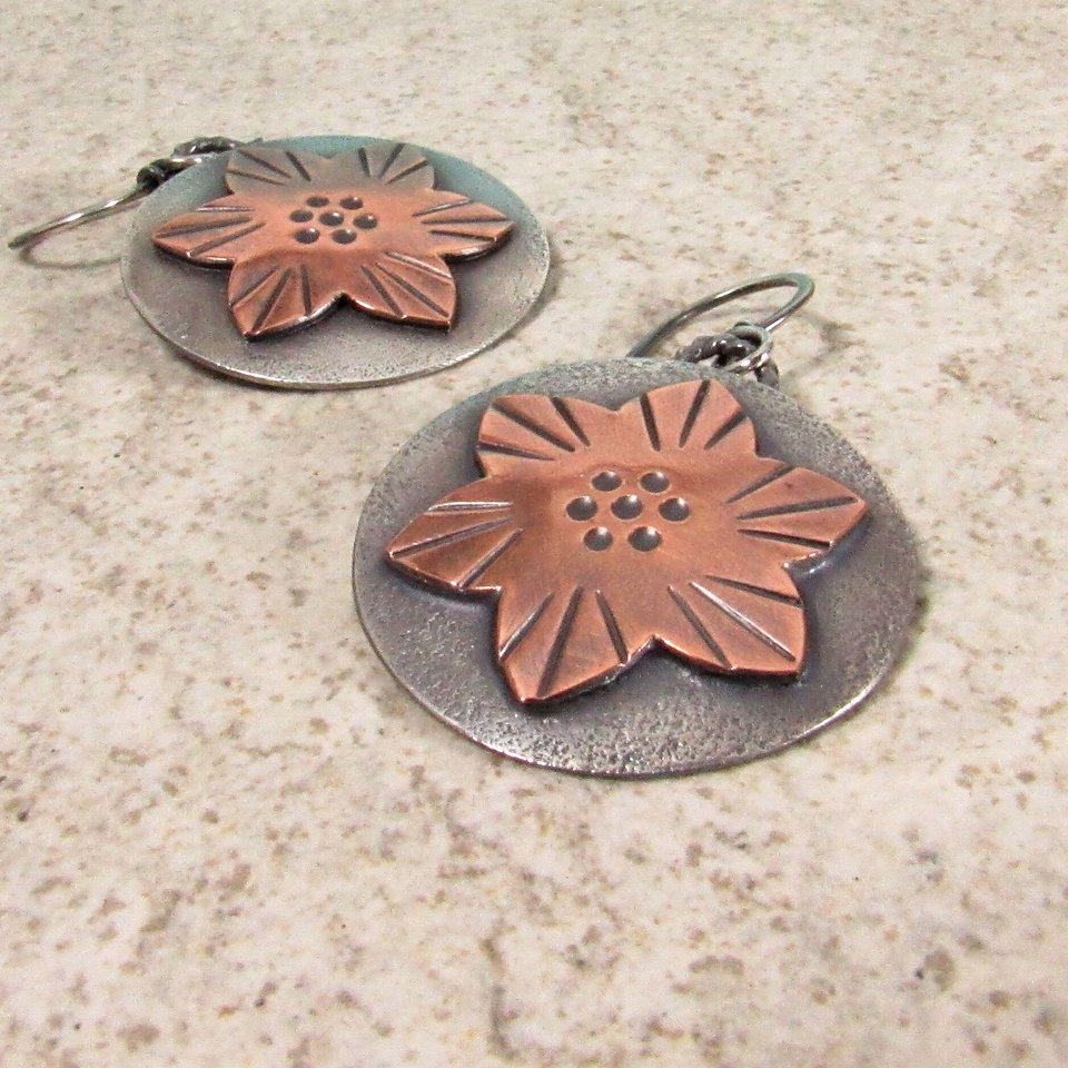 Mixed metal earrings feature a flower design in copper and sterling silver. They are a layered design, lightweight and measure 1.5" tall and 1" wide. The earrings are handcrafted and exceptional quality. Displayed on sand color background.