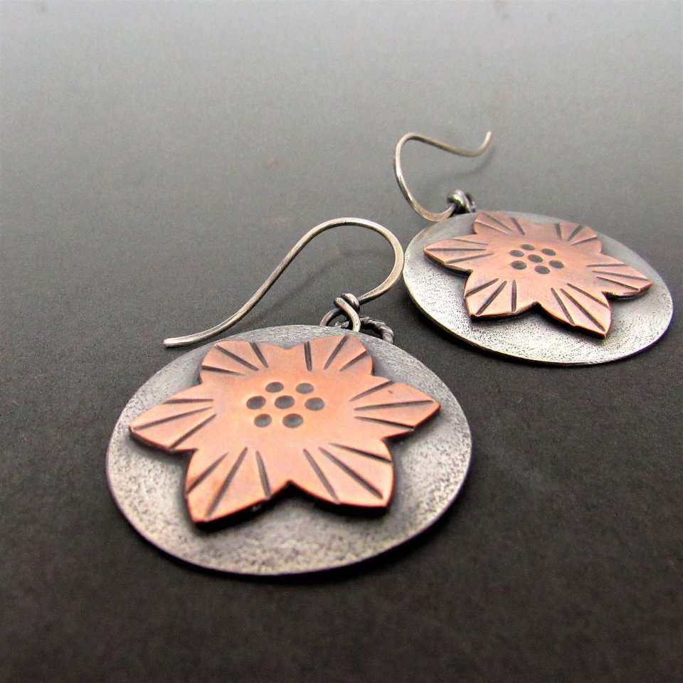 Handcrafted mixed metal star flower earrings in copper and argentium sterling silver. The copper flower sits atop the sterling silver disk. background. Argentium sterling silver ear wires complete these. Large enough to be noticed and light enough at a very comfortable weight for most at just over 5 grams each. Comfortable weight at 5 grams each. 1.5” long and 1” wide. Displayed against a dark background.1” wide