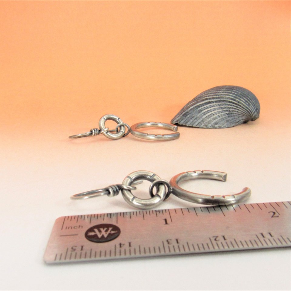 Bold and simple contemporary sterling silver earrings crafted from Argentium have lovely swingy movement. The double open hoop design allows for them to articulate. Lightweight and comfortable  at only 5 grams per earring. 1.75” long and 7/8” at widest point. Displayed against ruler on orange paper with other earring in background with a black shell