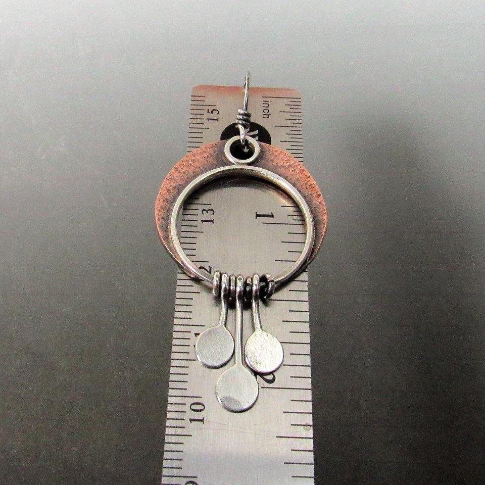 Copper and silver crescent moon dangle earrings with a circle design and three silver discs hanging from the bottom. The earrings are measured against a ruler, showing they are about 2 inch long.