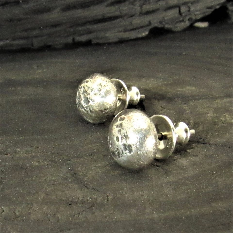 A pair of silver stud earrings with a rustic textured finish. The earrings are round and have a slightly irregular shape. Photo against dark background.