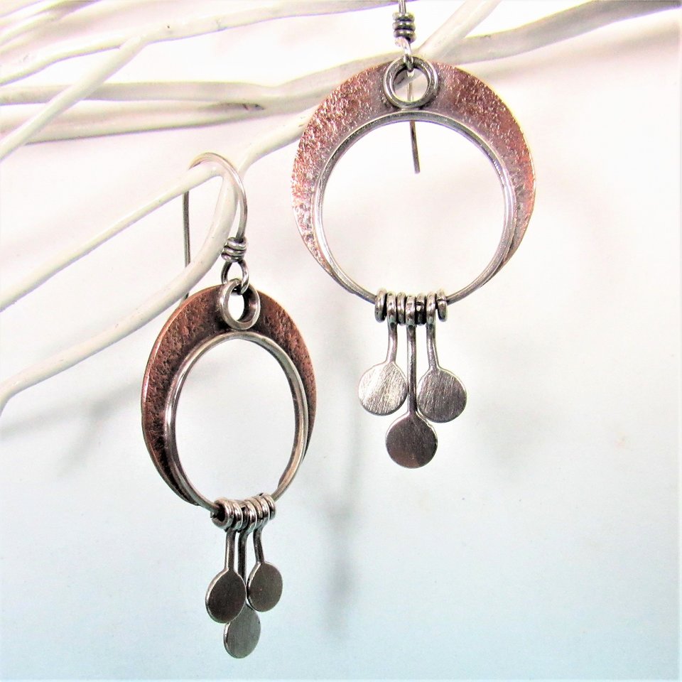 A lovely pair of mixed metal crescent moon earrings with moving paddles hanging from the crescent. Argentium silver and copper, 2 inch long, 1 inch wide. Swingy movement and lightweight. Perfect for dancing all night. Oxidized, polished, lovely glow. Displayed against white twigs on blue paper.