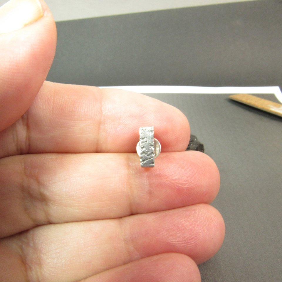 A silver textured bar earring with a post and butterfly back, displayed in hand.