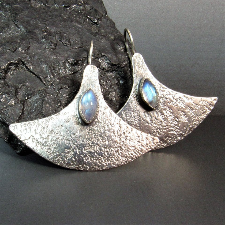 Large, futuristic argentium sterling silver and rainbow moonstone earrings with a touch of rusticity. Unusual and unique earrings that are modern with hints of the past. 1.5 inches tall x 1.75 inches wide. Displayed against rock, dark background.