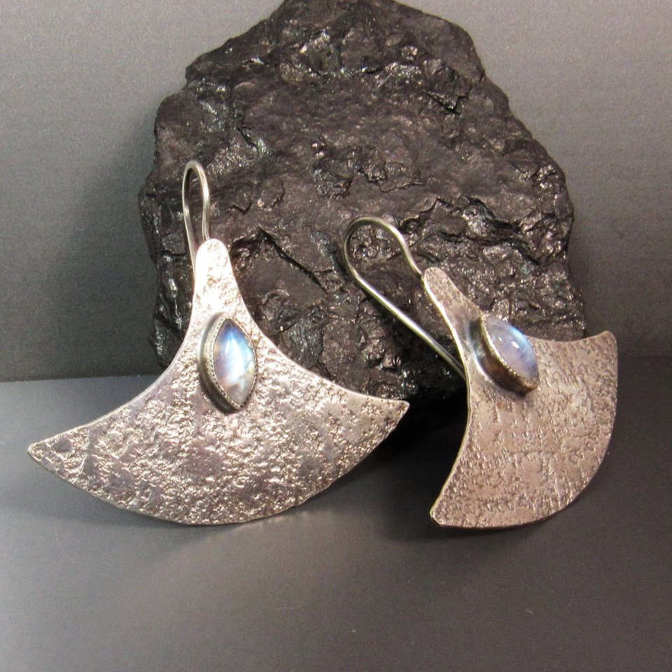 Large, futuristic argentium sterling silver and rainbow moonstone earrings with a touch of rusticity. Unusual and unique earrings that are modern with hints of the past. 1.5 inches tall x 1.75 inches wide. Displayed against rock, dark background.