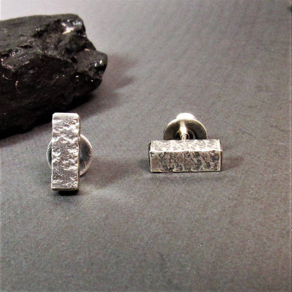 Front view of a pair of sterling silver stud earrings with a textured rectangular design. The earrings have a post back. Displayed on grey background with dark rock.