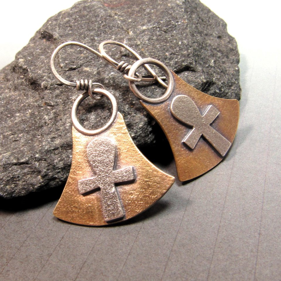 Mixed metal, bronze and silver ankh earrings. Body of earring shaped similar to the egyptian lotus motif with ankh on the front, suspended by silver ring and ear wires. 1.5" tall and 1" wide at widest point.  Displayed against black rock on grey background.