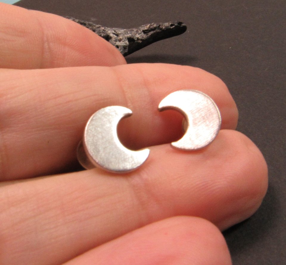 A pair of silver crescent moon earrings. The earrings are small and delicate, and they are perfect for adding a touch of whimsy to any outfit. Photographed in hand.