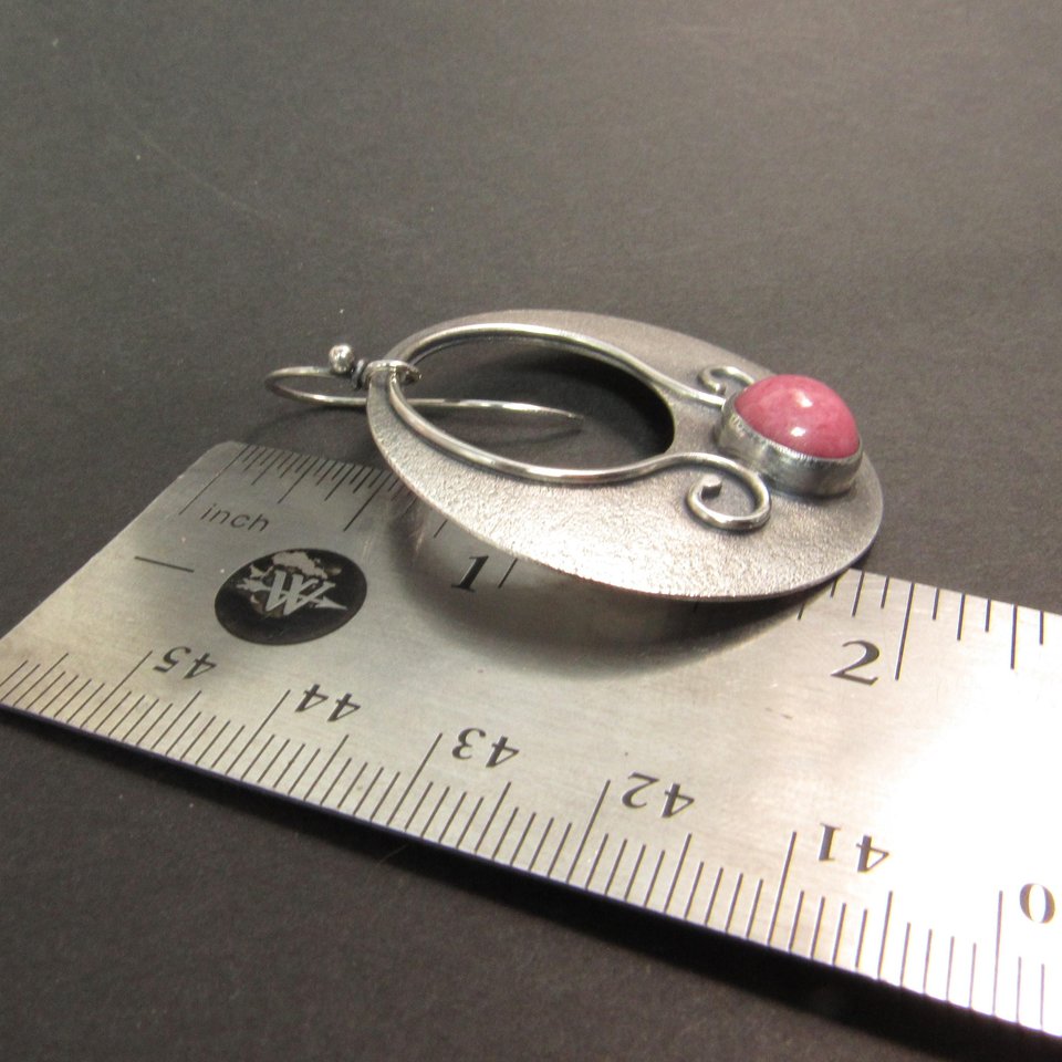 Large sterling silver earrings with earthy deep pink rhodonite cabochons, textured surface and  swirling SS wire embellishments. Oxidized to add depth and drama. Earrings measure apx 1.75” tall and a touch over 1.5” wide. Displayed against ruleron dark background.