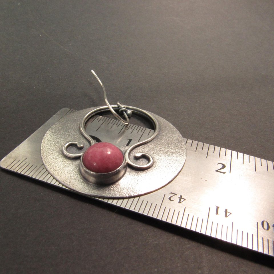 Large sterling silver earrings with earthy deep pink rhodonite cabochons, textured surface and  swirling SS wire embellishments. Oxidized to add depth and drama. Earrings measure apx 1.75” tall and a touch over 1.5” wide. Displayed against ruler on dark background.