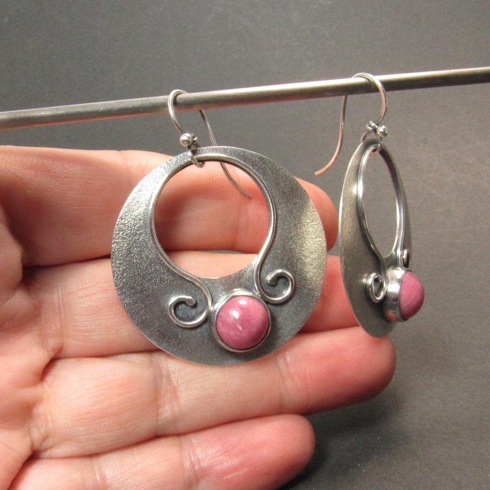 Large sterling silver earrings with earthy deep pink rhodonite cabochons, textured surface and  swirling SS wire embellishments. Oxidized to add depth and drama. Earrings measure apx 1.75” tall and a touch over 1.5” wide. Displayed against hand on dark background.