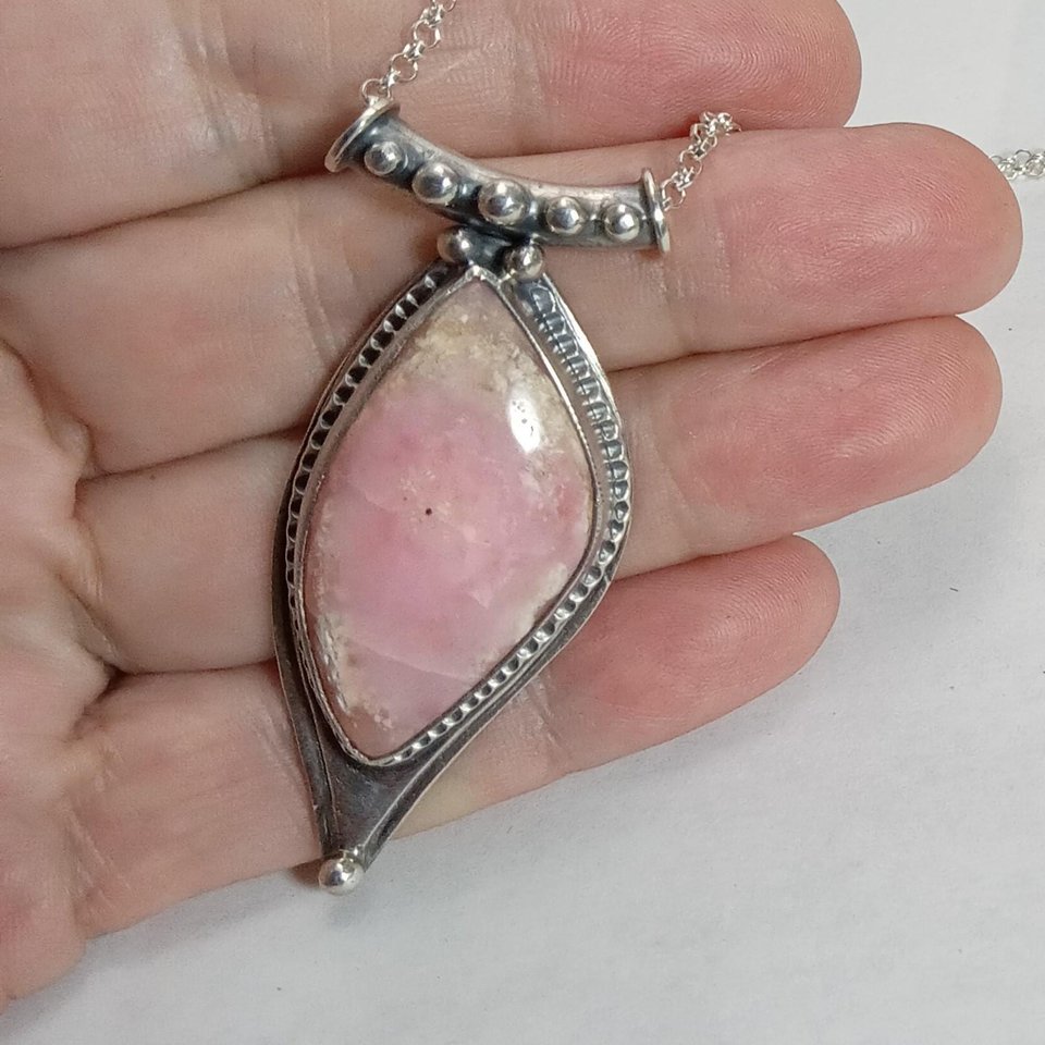 Soft elegance meets artisan craftsmanship in this one of a kind handcrafted pendant featuring a beautifully pastel pink opal set in textured Argentium® sterling silver. The unique, petal-shaped stone in a petal shaped setting radiates gentle warmth in a soothing blush hue—perfect for those who adore subtle statement pieces with soulful charm. 2.25" long x 7/8" wide.